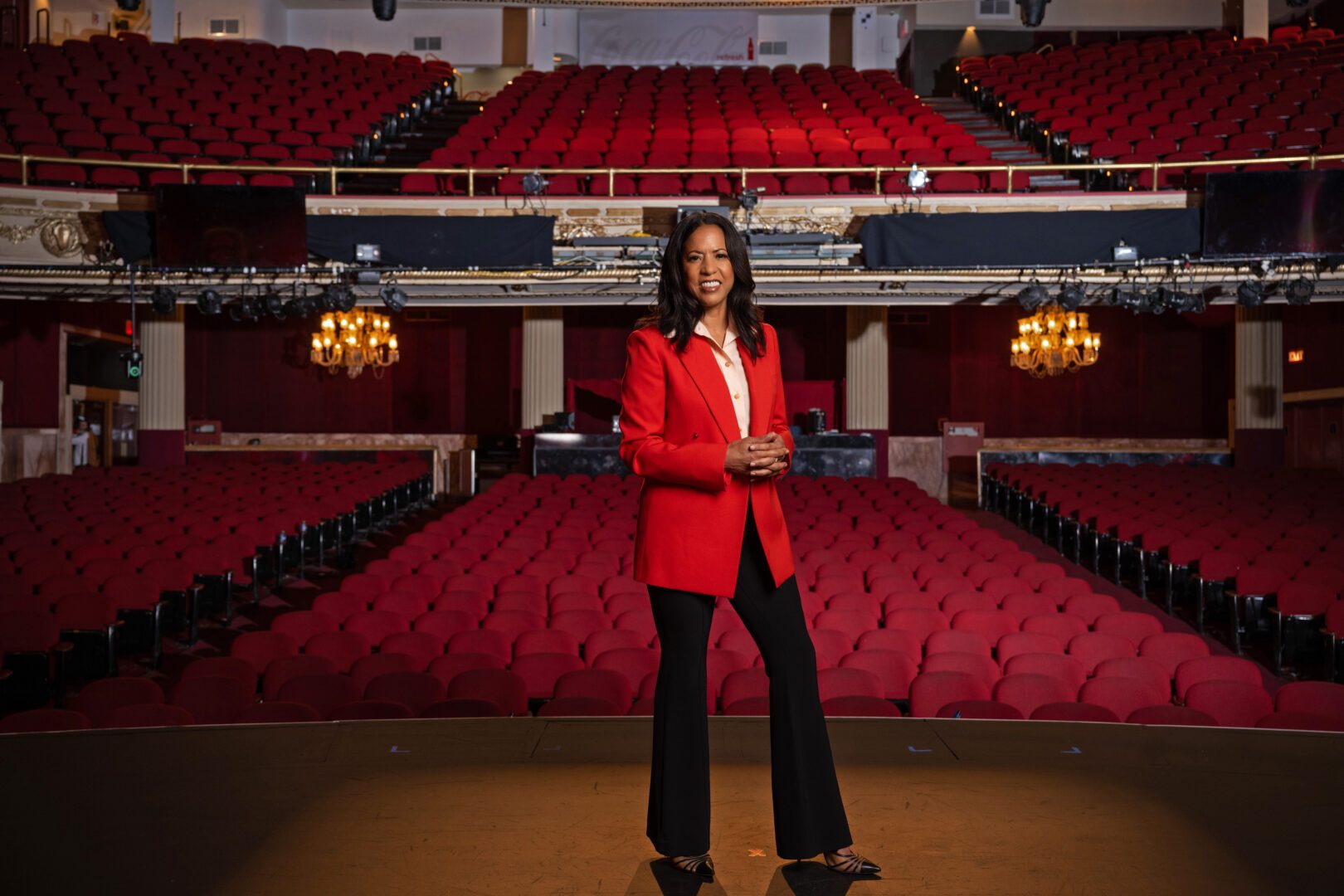 A Black Woman with long hair with a red blazer and black pants and standing on the The Apollo's historic theater stage