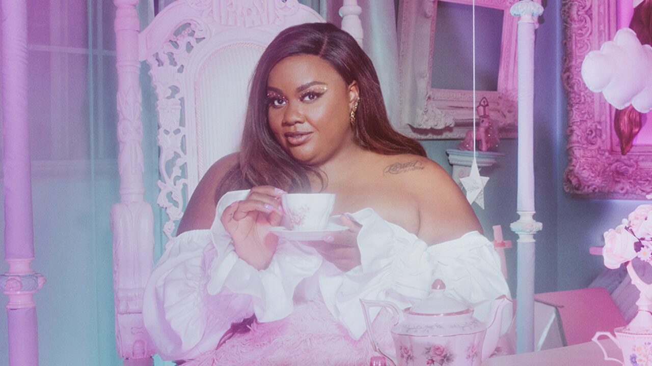 Nicole Byer - a Black woman with long hair in a light pink dress holding a cup of tea